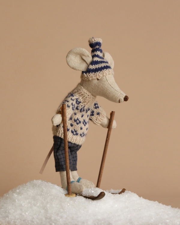 A Maileg Winter Mouse with Ski Set, Dad - Blue is positioned atop a small snow-covered hill, dressed in a knitted sweater and hat, standing on skis with ski accessories and ready for winter adventures against a neutral background.