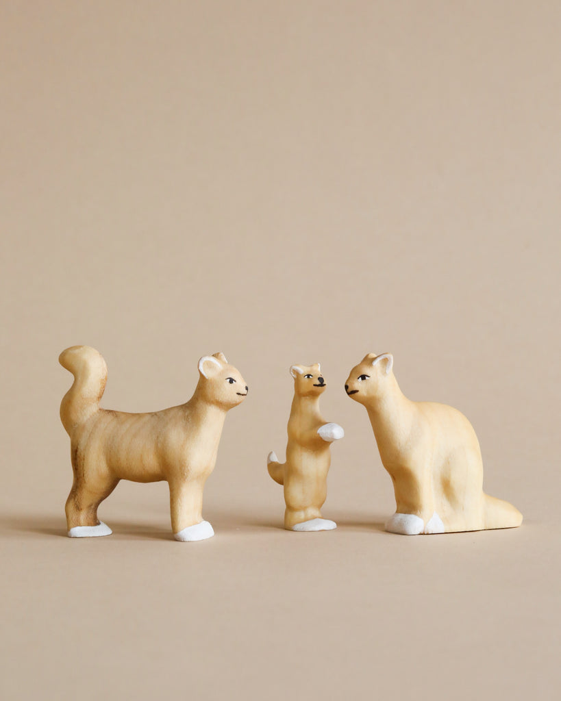 The Handmade Wooden Cat Family features three handcrafted wooden cat figurines displayed against a beige background. One cat stands on all four legs with its tail curled upward, another cat is sitting on its hind legs, and the third sits upright. Made from Romanian ash wood and finished with water-based paint, they boast white paws and a light brown color.