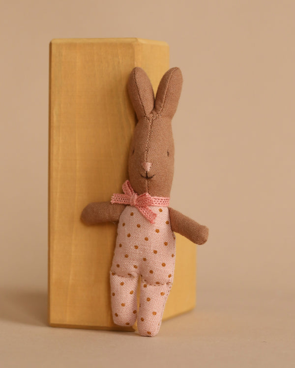 A Maileg My Rabbit - Dots, a plush brown bunny adorned with a pink bow and polka-dotted details, is propped against a wooden block against a neutral backdrop.