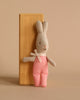 The Maileg My Rabbit - Red Check, a small stuffed bunny in red and white checkered overalls with a bow around its neck, is leaning against a wooden block on a beige background.