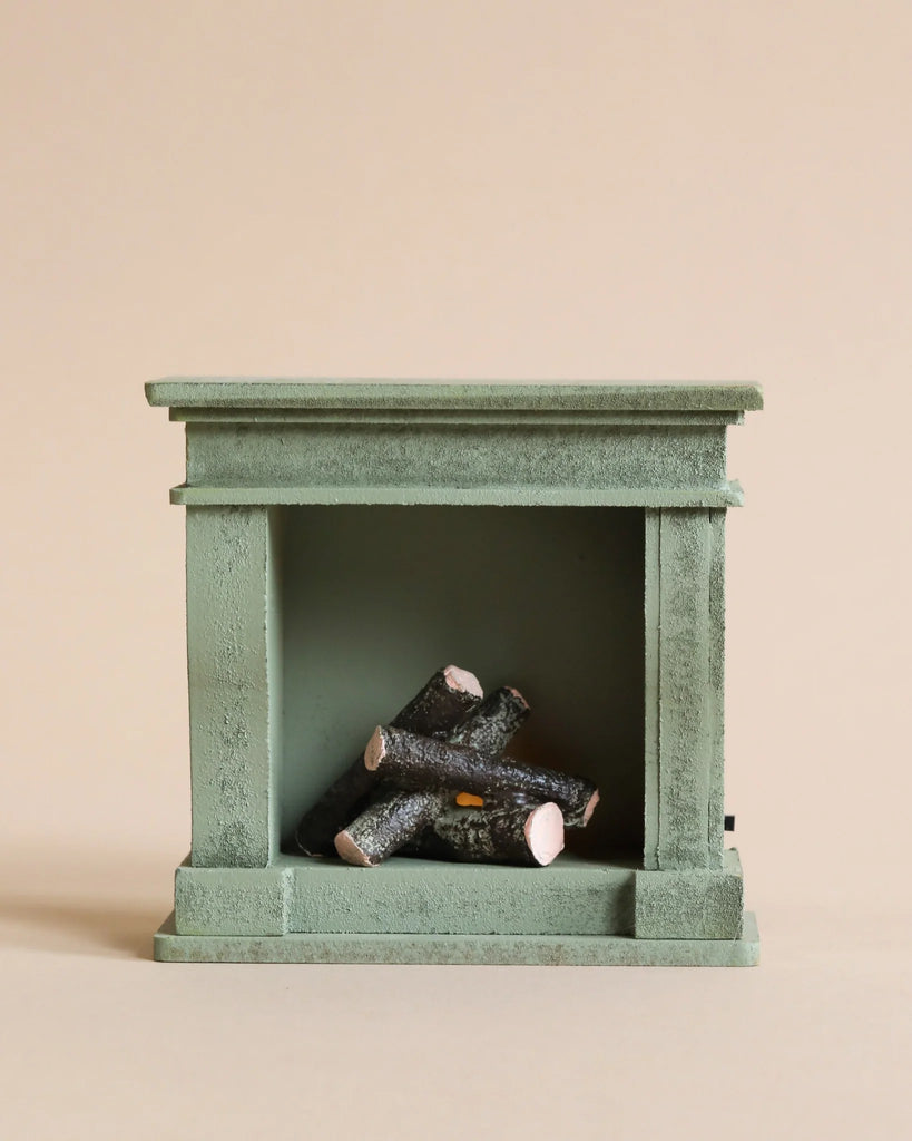 A miniature green fireplace, featuring a straightforward design with three small, realistic logs inside, is set against a plain light beige background. Reminiscent of a cozy corner in a Maileg Castle, this piece enhances the minimalistic aesthetic and complements the elegance of the Maileg Royal Tea Party.