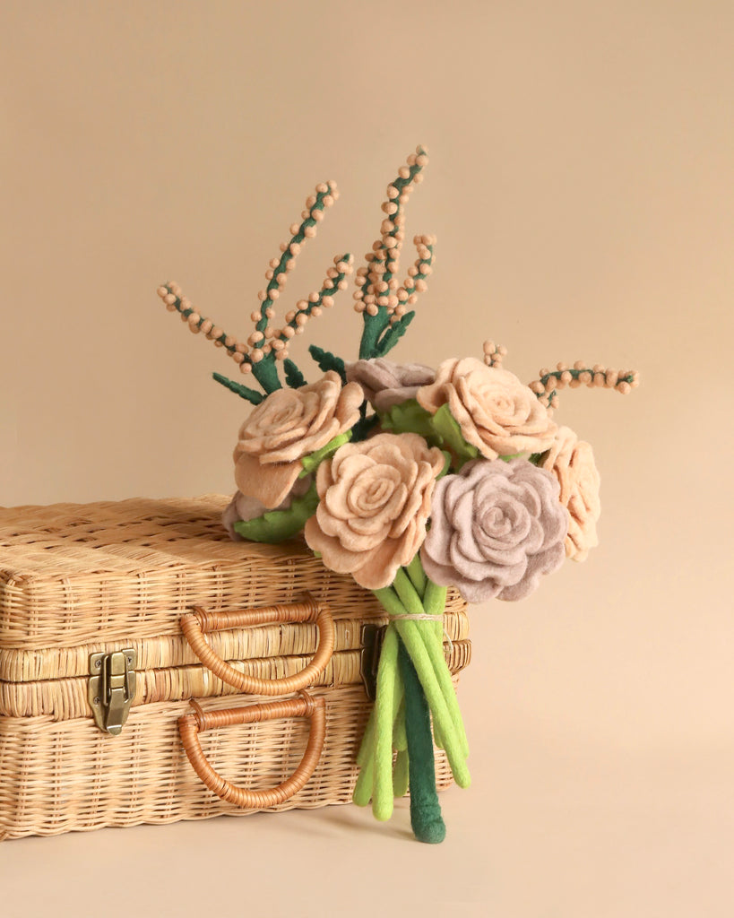 A bouquet of eco-friendly handcrafted Blooming Rose felt flowers in shades of brown, pink, and lavender rests on a woven wicker suitcase with a beige background.