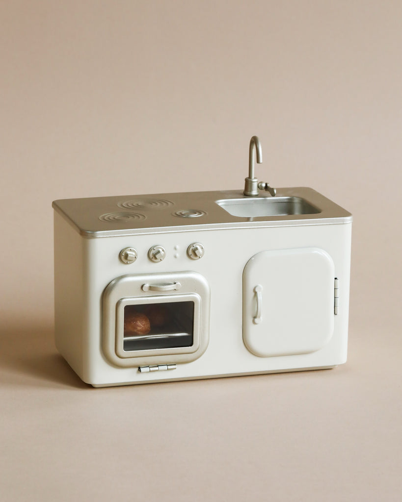 A Maileg Mini Kitchen miniature play kitchen set in neutral tones, featuring a sink, stove, and oven with freshly baked buns inside on a light beige background.