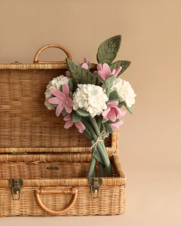 A wicker picnic basket with its lid slightly open, revealing a bouquet of Felt Flower Bouquet - White Hydrangea with green leaves, set against a neutral background.