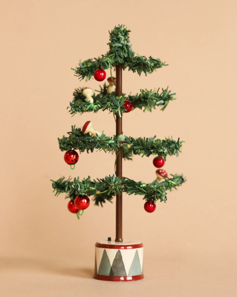 A small, minimalist Christmas tree from the Maileg Christmas Set features sparse green branches adorned with a few red and white ornaments, standing on a decorative base with geometric patterns reminiscent of a gingerbread house, set against a plain beige background.