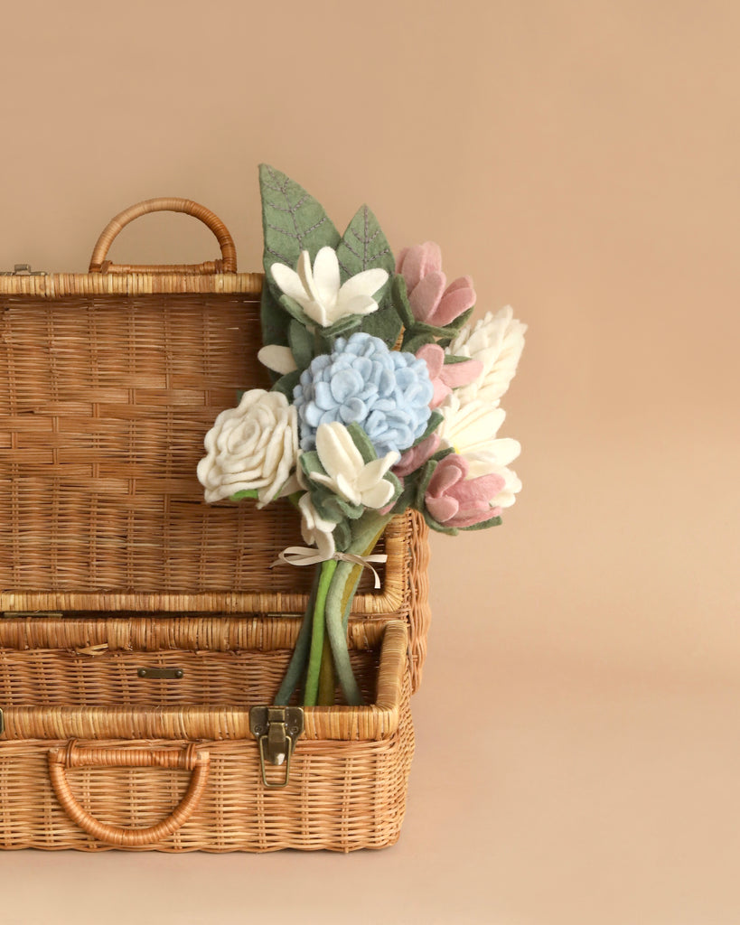 A vintage wicker picnic basket with a handle and the Felt Flower Bouquet - Muted Blooms peeking out against a muted beige background.