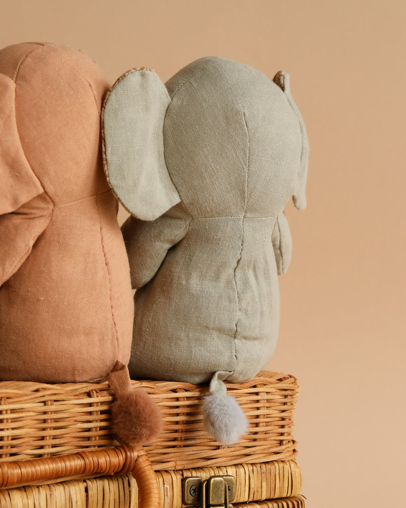 Sentence with Product Name: Two Maileg Small Elephant toys, one gray and one brown, sitting on a wicker basket with a soft beige background. Both safari friends have their backs to the viewer, displaying large floppy ears and tiny tails.