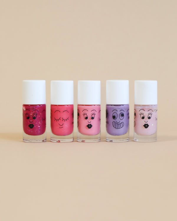 Five bottles of Nailmatic water-based nail polishes with cartoon faces on a beige background, ranging in colors from deep red to soft pink, each showing different facial expressions.