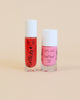 Two bottles of Nailmatic water-based nail polish with cute faces drawn on them, one pink and one red, against a soft peach background.