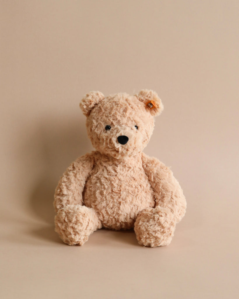 A fuzzy, light brown Steiff Jimmy Teddy Bear sitting upright against a plain, beige background. The bear has black eyes, a shiny nose, and a gently smiling expression.