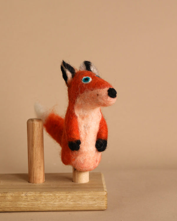 The Fox Finger Puppet, handcrafted from wool felt featuring an orange body, white belly, and black details, is elegantly displayed on a wooden stand against a simple beige background. Its blue eyes add a playful and whimsical touch, making it perfect for imaginative storytelling.