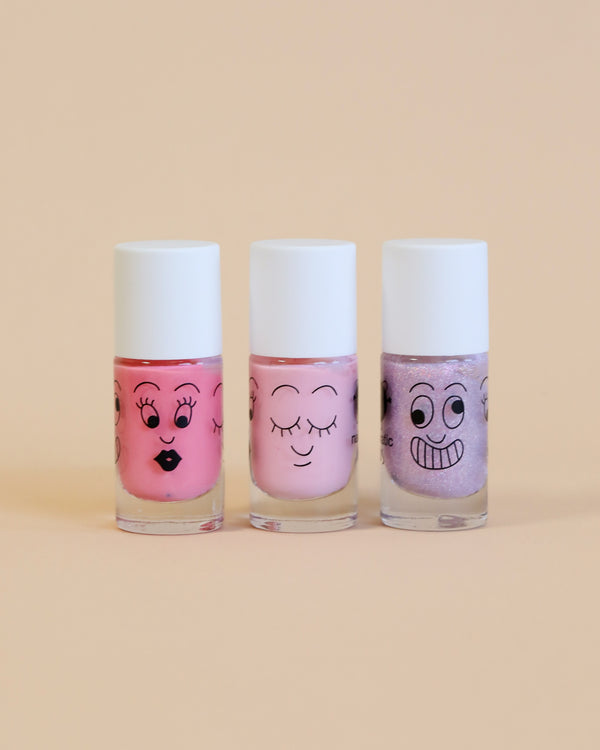 Three bottles of Nailmatic water-based nail polishes with cartoon faces drawn on them, in shades of pink and purple, against a pale yellow background.