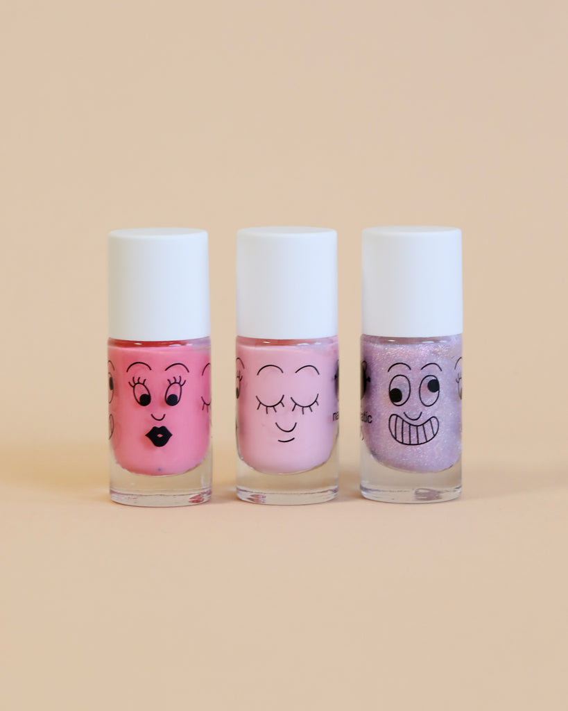 Three bottles of Nailmatic water-based nail polishes with cartoon faces drawn on them, in shades of pink and purple, against a pale yellow background.