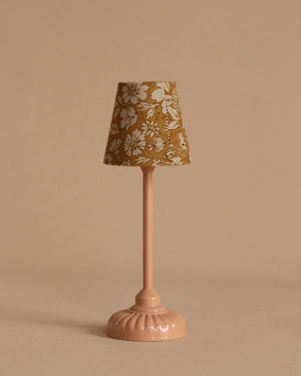 A small, pink table lamp with a floral-patterned lampshade stands against a beige background, casting a soft tone light. The shade showcases white flowers on a mustard yellow backdrop, reminiscent of the design found on the Maileg Mouse Size Vintage Floor Lamp in Dark Powder.