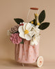 A small, pink polka-dotted Felt Flower Trolley Basket sits on a metal cart with two white wheels. The basket contains a vibrant arrangement of handmade felt flowers in white, pink, purple, and yellow colors, along with green felt leaves. Crafted with care and commitment to fair wages, it rests against a plain light tan background.