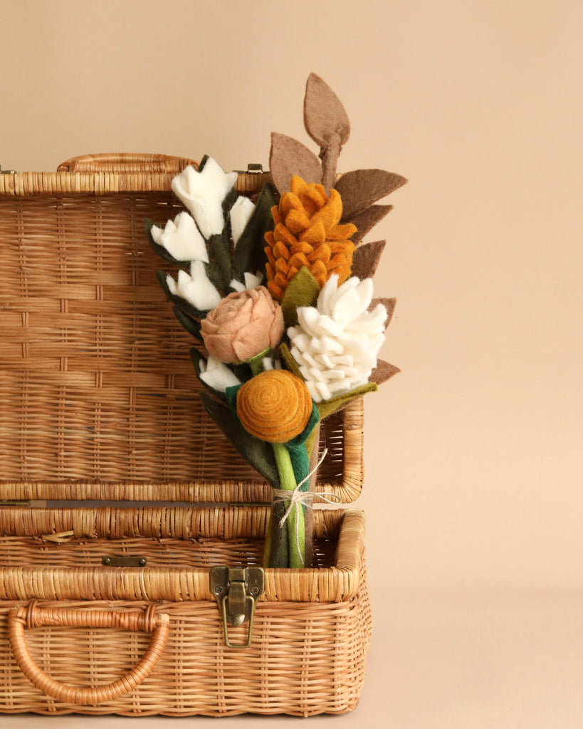 A small Felt Flower Bouquet - Golden Hour, including white, orange, and peach blooms, arranged with green and brown felt leaves, placed inside an open wicker suitcase against a beige background.