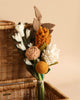 A Felt Flower Bouquet - Golden Hour is displayed against a wicker basket. The handmade flowers exhibit various textures and forms, creating a visually engaging arrangement on a beige background.