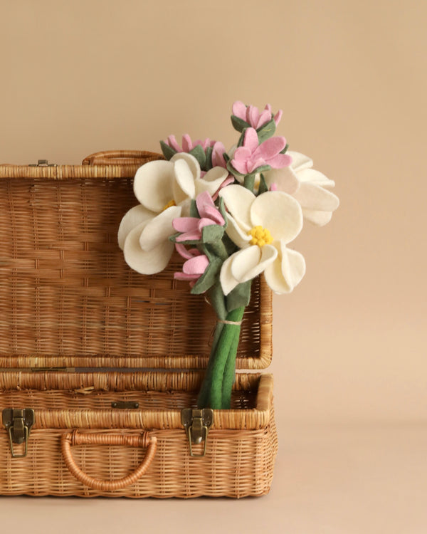 A wicker suitcase is open with a Felt Flower Bouquet - Magnolia inside. The bouquet includes large white flowers with yellow centers and smaller pink flowers, all with green stems. The background is a plain beige, complementing the natural tones of the suitcase and flowers.