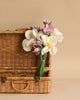 A wicker suitcase is open with a Felt Flower Bouquet - Magnolia inside. The bouquet includes large white flowers with yellow centers and smaller pink flowers, all with green stems. The background is a plain beige, complementing the natural tones of the suitcase and flowers.