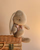 A Maileg Medium Plush Bunny in dusty brown, featuring floppy ears and crafted from soft fabric with polyester fillings, is adorned with a pink ribbon as it sits on a wicker basket against a beige background.