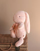 A Maileg Medium Plush Bunny - Powder, crafted from soft fabric and recycled polyester, sits gracefully on a woven basket. Its long floppy ear and delicate ribbon around its neck enhance its charm against a neutral background.