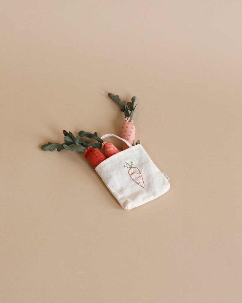 A small beige cloth bag with a carrot illustration, containing several stuffed fabric carrots with green leaves for imaginative play, is placed on a light brown surface.