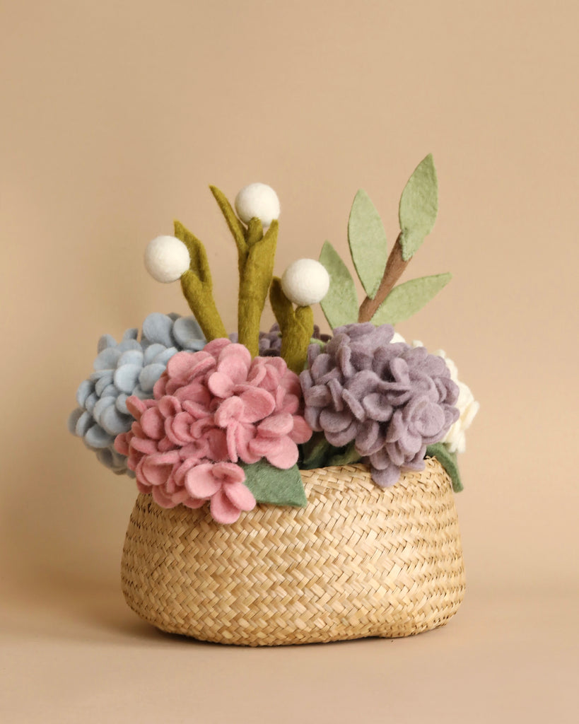 A Basket of Felt Hydrangea Flowers filled with colorful, handmade felt flowers, including blue, pink, and purple blooms, along with green felt leaves and white globe-like buds, is set against a solid beige background. The eco-friendly arrangement adds a charming and whimsical touch.