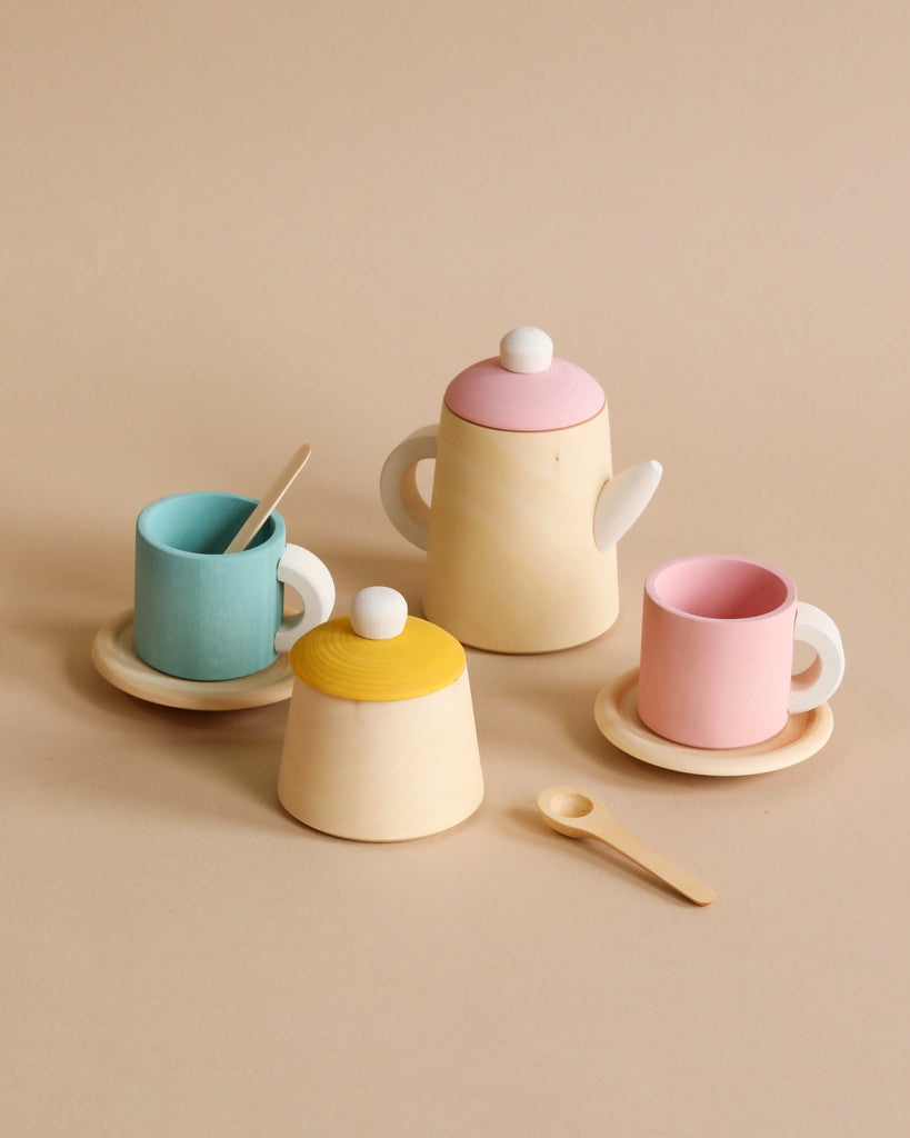 A Raduga Grez Handmade Wooden Tea Set featuring a pink-lidded teapot, a yellow-lidded sugar bowl, blue and pink cups with matching saucers and spoons, all crafted with non-toxic paint on a neutral beige background. Designed with simple, smooth lines and bright pastel colors, it’s perfect for pretend play.