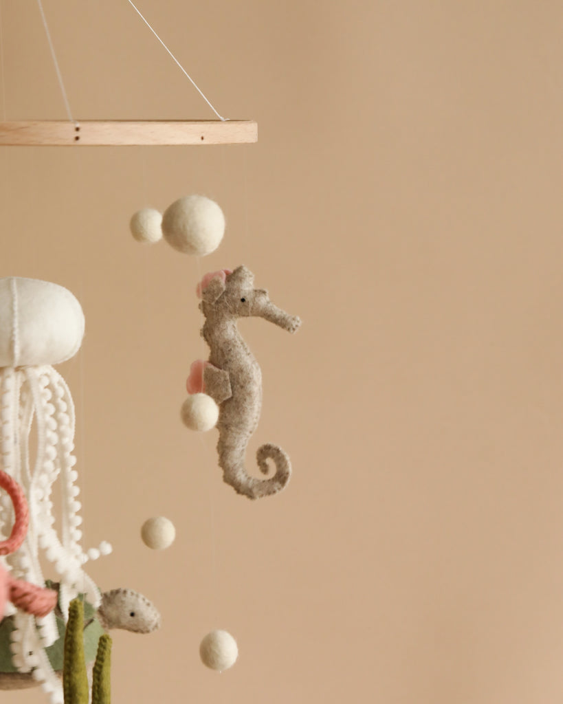 A close-up of a Handmade Mobile - On The Reef - Final Sale features a cute felt seahorse with soft pink accents, handmade in Europe, suspended alongside other sea-themed elements like an octopus, small fish, and large white felt balls. Perfect for nursery decoration, the background is a neutral beige color.