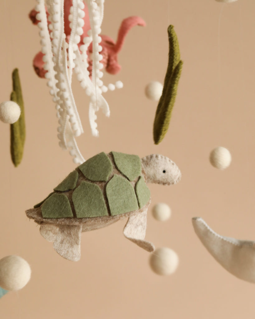 A close-up of a whimsical felt mobile, **Handmade Mobile - On The Reef - Final Sale**, featuring a green sea turtle with a patchwork shell, white fluffy balls, and seaweed-like felt pieces hanging from strings. Perfect for nursery decoration and visual development, the soft beige background adds to the overall gentle and soothing feel.