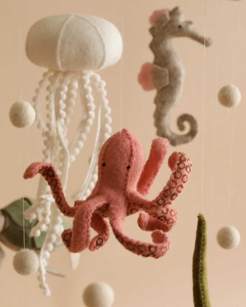 A Handmade Mobile - On The Reef - Final Sale, handmade in Europe, featuring felt sea creatures: a white jellyfish with trailing tentacles, a pink octopus with detailed tentacles, a gray seahorse with a pink fin, and several white felt balls hanging on strings—perfect for visual development and nursery decoration.