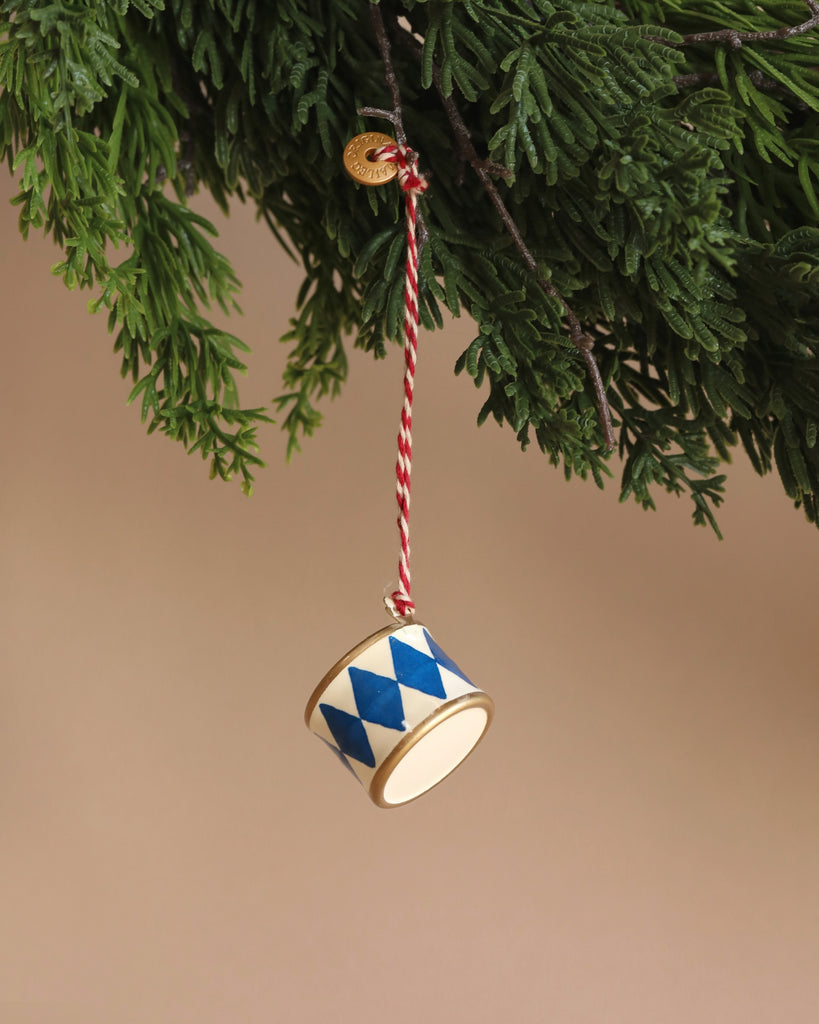 A Maileg Metal Ornament, Small Drum in dark blue with blue diamond patterns and delicate handpainted details hangs from a branch of a coniferous tree using a red-and-white striped string, set against a soft, neutral background.