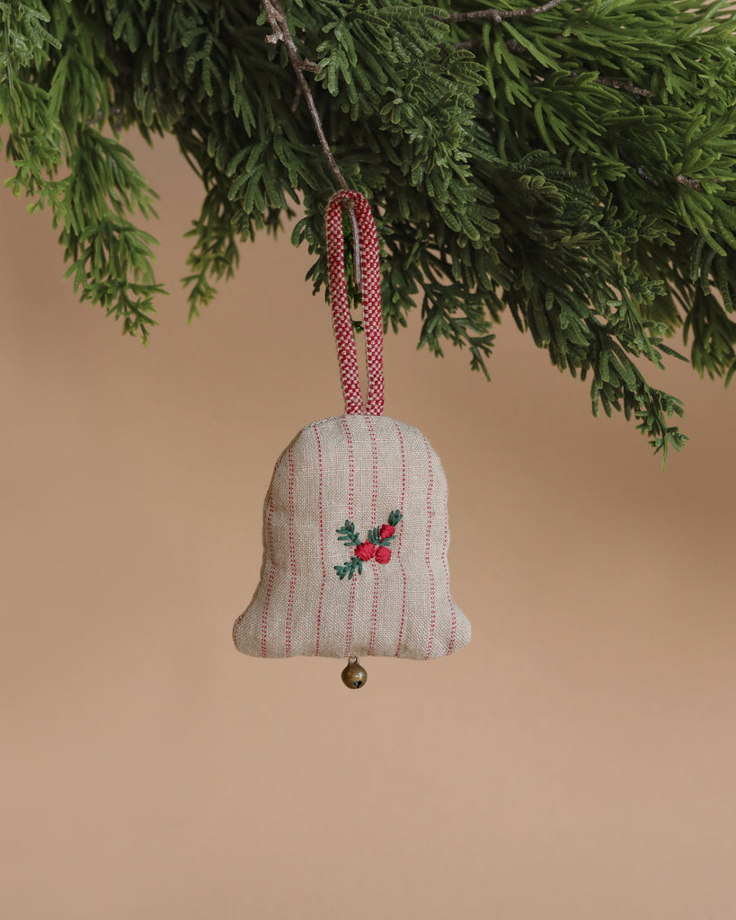 The Maileg Ornament - Small Bell, with its handmade fabric construction and red embroidered flower design, adorns a Christmas tree branch. Its features include a red plaid loop and a small golden chime at the bottom, capturing Maileg's enchanting style against a soft beige backdrop.