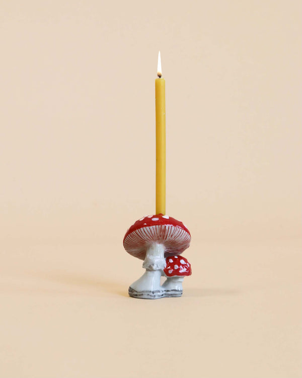 A lit yellow candle standing in a whimsical, heirloom-quality Mushroom Cake Topper shaped like a red and white spotted mushroom against a pale beige background.