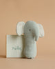 A small, light gray Maileg Lullaby Friend Rattles, Elephant - Light Dusty Blue with large white ears stands in front of a beige box. The word "Maileg" is written on the box in green cursive lettering. This delightful elephant doubles as a soft fabric rattle, perfect for little hands. The background is a plain, light brown shade.