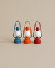 Three Maileg Lanterns are placed side by side against a neutral background. Each non-rechargeable lantern features a white globe and comes in a different color: blue, orange, and red. The lanterns are perfectly aligned, creating a visually appealing arrangement.