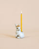 A whimsical hand painted porcelain candle holder shaped like a Royal White Rabbit Cake Topper adorned with a golden crown, holding a lit yellow candle against a soft beige background.