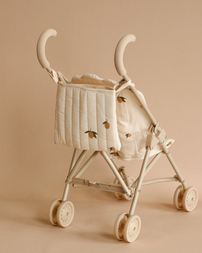 A Konges Sloejd Doll Stroller - Lemon with a padded backrest featuring a subtle pattern of small yellow birds. The stroller has smooth, double white wheels and curved handles, along with a secure harness. The background is a solid, light brown color.