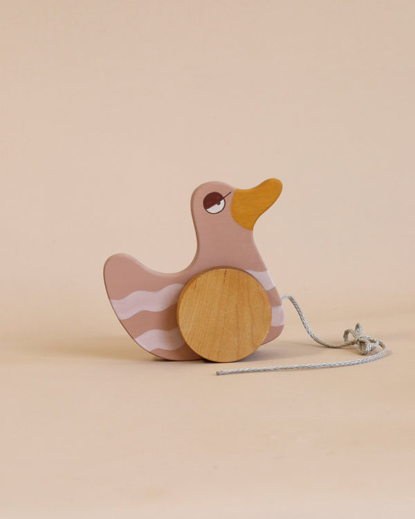 A Duck Pull Toy - Pink with a pink and white body, an orange beak, and wooden wheels, standing on a beige background with a pull-string attached is crafted from sustainably harvested birch wood.