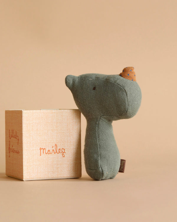 A small, plush, green toy bear with a tiny brown hat rests beside a beige, textured gift box with "Maileg Lullaby Friend Rattles, Rhino - Pine" written on it in orange. The background is a muted beige color, enhancing the cozy feel of the scene.