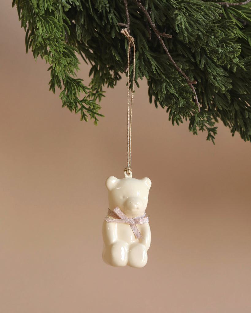 The Maileg Metal Ornament, Teddy Bear in Off White/Rose, with handpainted details and a pink ribbon around its neck, hangs from a green branch against a beige background.