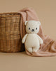 A Cuddle + Kind Baby Polar Bear made from heirloom-quality natural Peruvian cotton yarn with a happy expression sits against a woven basket, draped with a pink knit blanket, on a beige background. The scene evokes a sense of warmth and coziness.