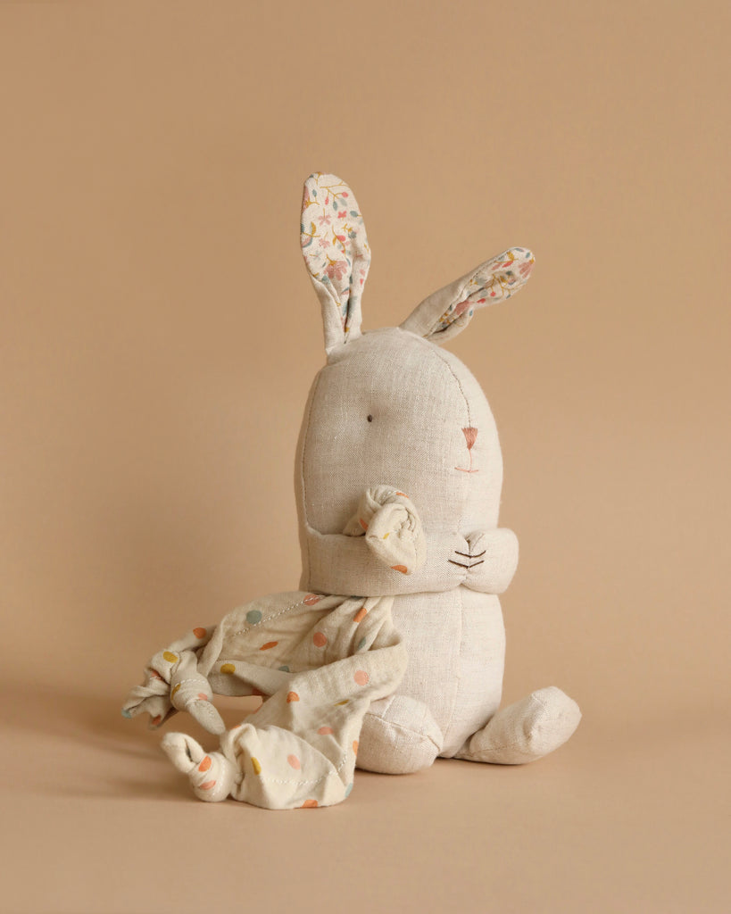 A Maileg Lullaby Friends - Bunny - Nature (Plays Music) with floral-patterned inner ears and a minimalist face holds a small, colorful polka-dotted bunny cuddle cloth. The bunny sits upright against a solid beige background, creating a simple and cozy atmosphere.