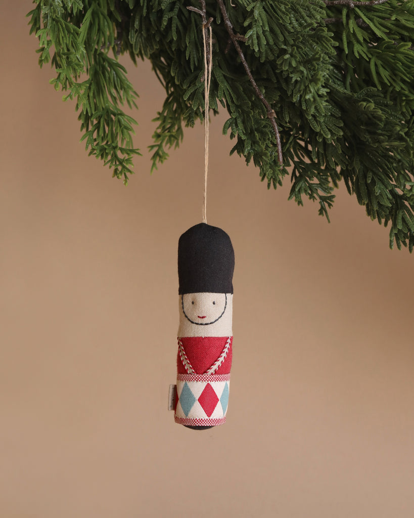 The Maileg Guard Ornament - Red, a handmade decoration in the shape of a toy soldier, hangs on the Christmas tree. The ornament features a black hat and a red and blue uniform with crisscross patterns and simple facial details, all set against a neutral tan background.