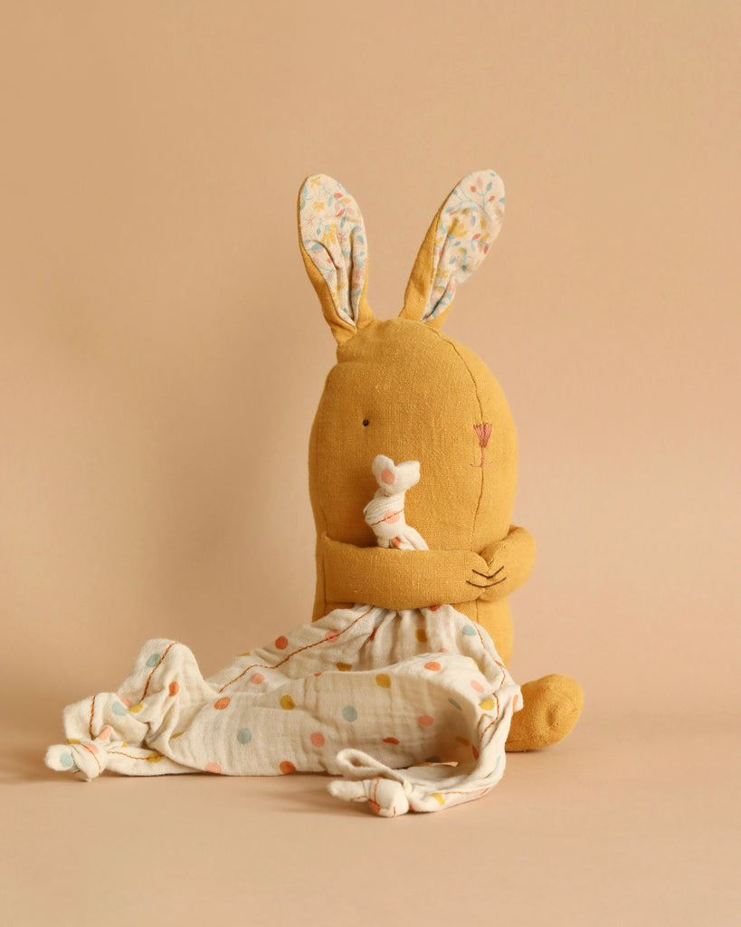 A Maileg Lullaby Friends - Bunny - Dusty Yellow (Plays Music) from the collection with floral-patterned inner ears sits against a beige background. It cuddles a small, gray stuffed mouse and is draped with polka-dotted fabric, evoking the serene notes of Mozart's lullaby in a warm, cozy, minimalist scene.