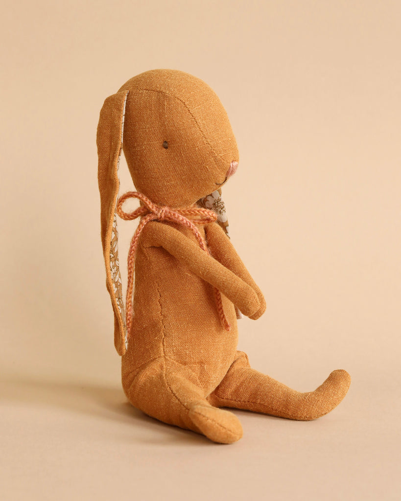 A Maileg Bunny - Ocher made from recycled polyester sits against a matching brown background. The brown fabric toy features long ears, small black eyes, and a stitched pink nose. It wears a knotted pink ribbon around its neck and has magnets in its hands. The bunny is in a seated, slightly side-facing position.