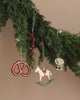 A garland of green pine branches adorned with the Maileg Peter's Christmas Ornament Set (2024), featuring a red and white candy cane, a red pretzel, a white rocking horse, and a small drum. A whimsical snowman ornament adds charm against the soft tan background.