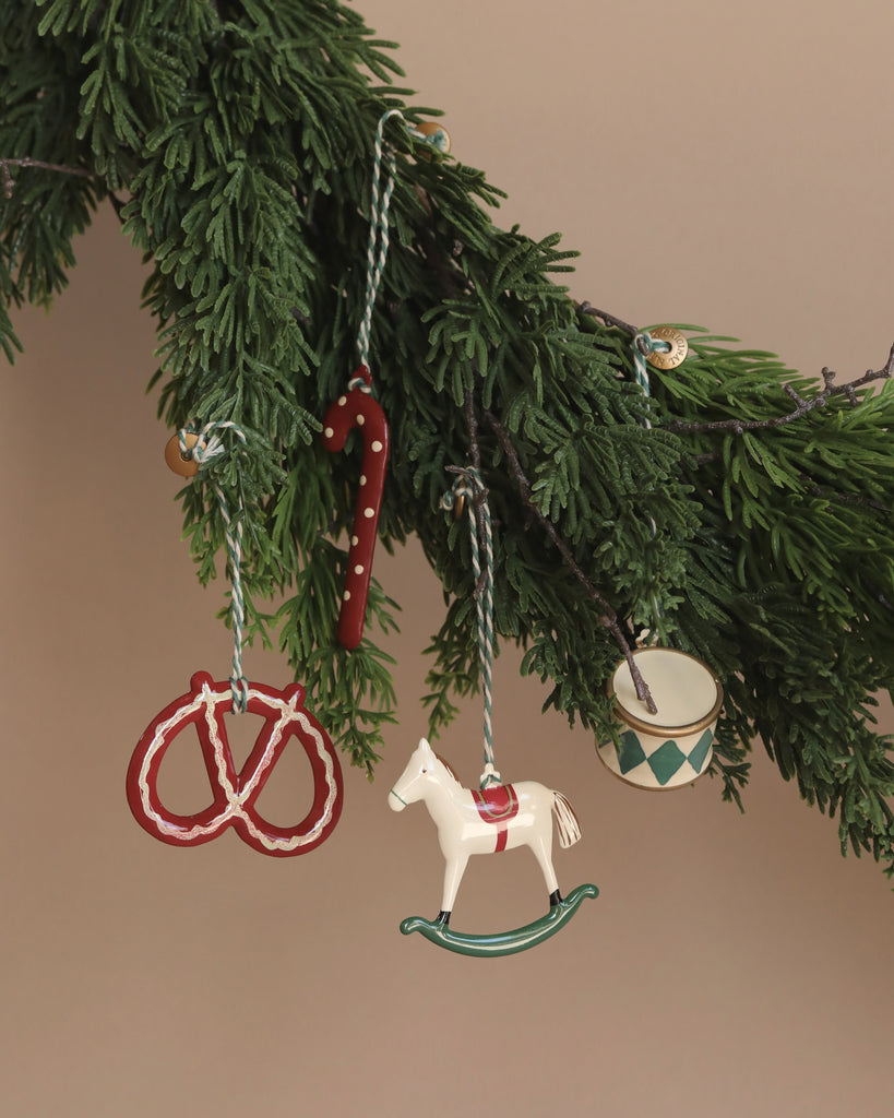 A garland of green pine branches adorned with the Maileg Peter's Christmas Ornament Set (2024), featuring a red and white candy cane, a red pretzel, a white rocking horse, and a small drum. A whimsical snowman ornament adds charm against the soft tan background.