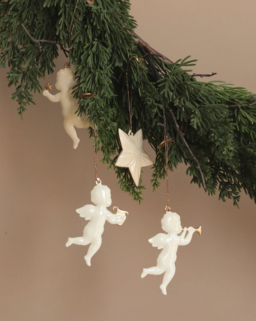 Green branches draped with the Maileg Holy Night Ornament Set, showcasing angelic figures with a trumpet and lyre and a star-shaped piece. Inspired by Dorte Mailil's sketches, this beautiful display pops against the beige wall, crafting a charming holiday scene.