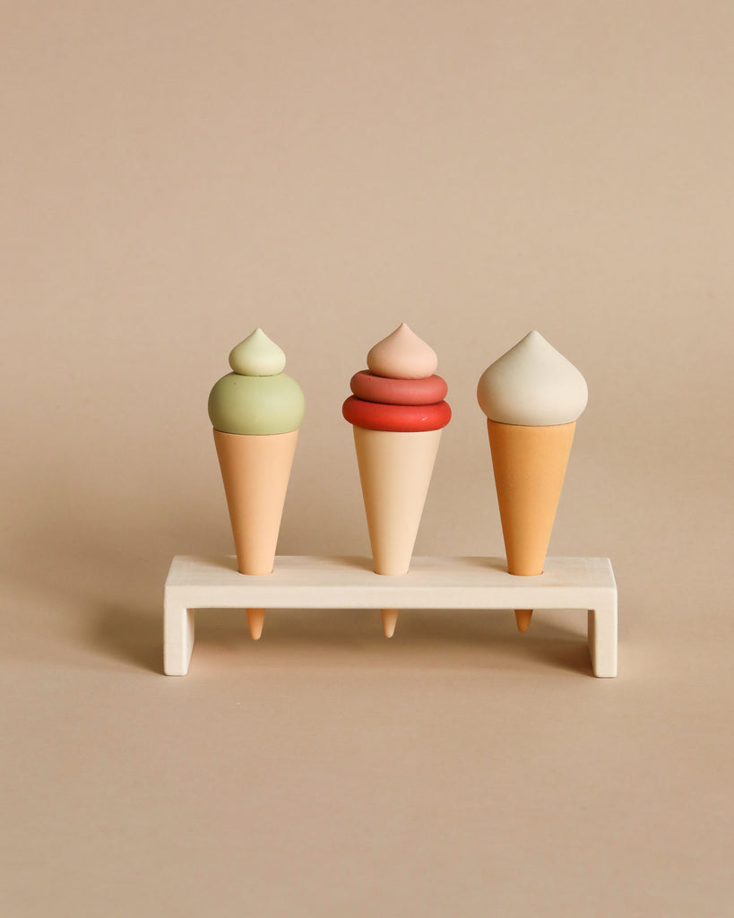 Three handmade ice cream cone toys with green, pink, and white swirls on top, displayed on a tiny wooden shelf against a neutral background.